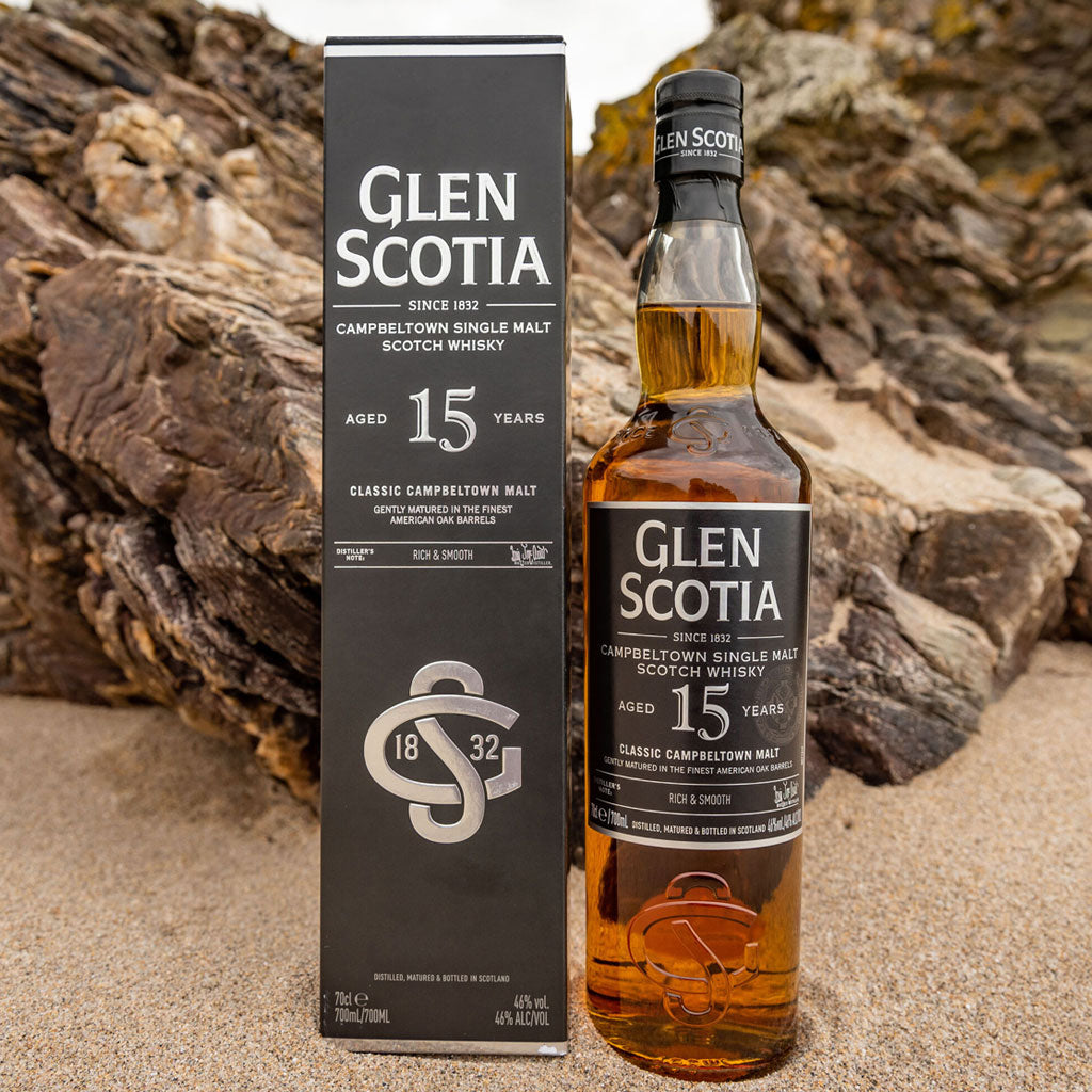 A 750ml bottle of Glen Scotia 15-year-old Campbeltown single malt scotch whisky is placed next to a black and white box with the same branding. The background has rocky terrain, and the box and bottle are securely positioned on sand.