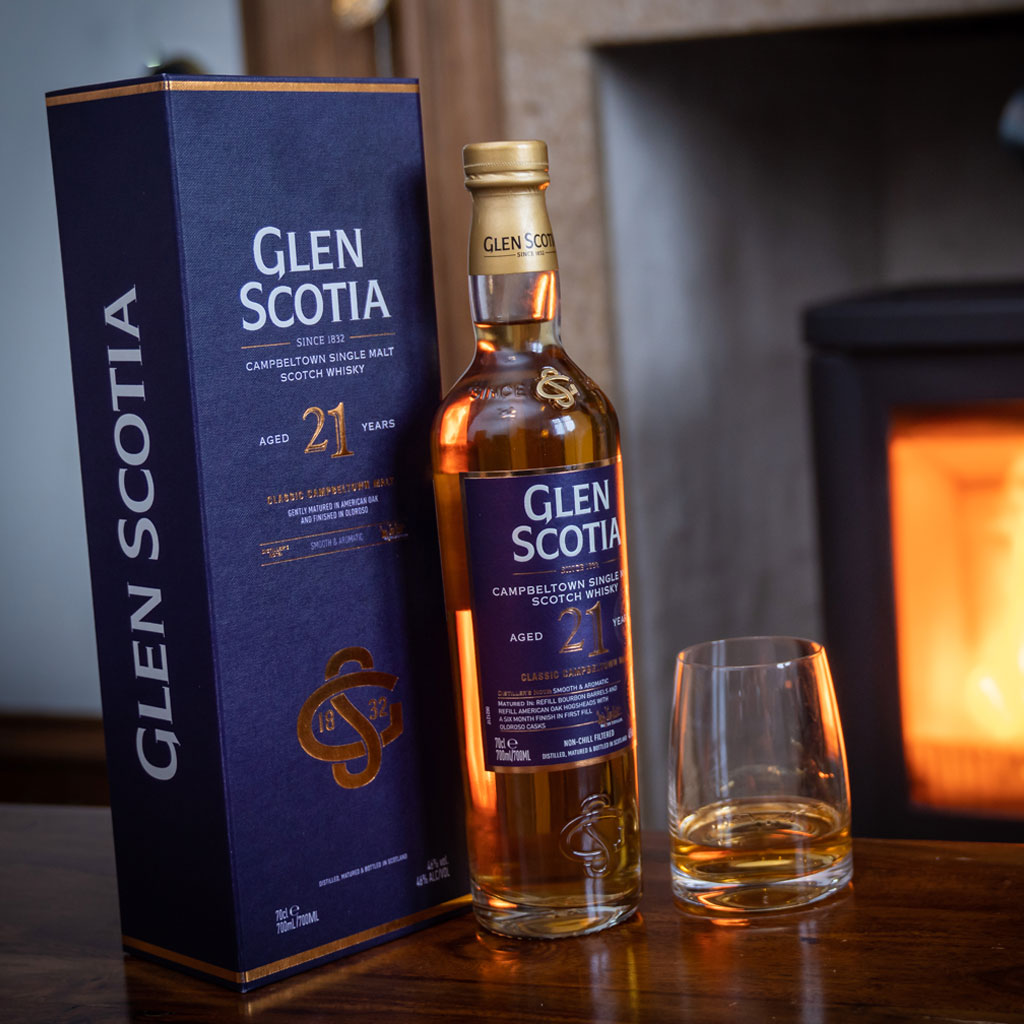 A bottle of Glen Scotia 21-year-old Campbeltown Single Malt Scotch Whisky is displayed next to its box packaging on a wooden surface. A glass with whisky is placed beside the bottle. In the background, there is a cozy fireplace.
