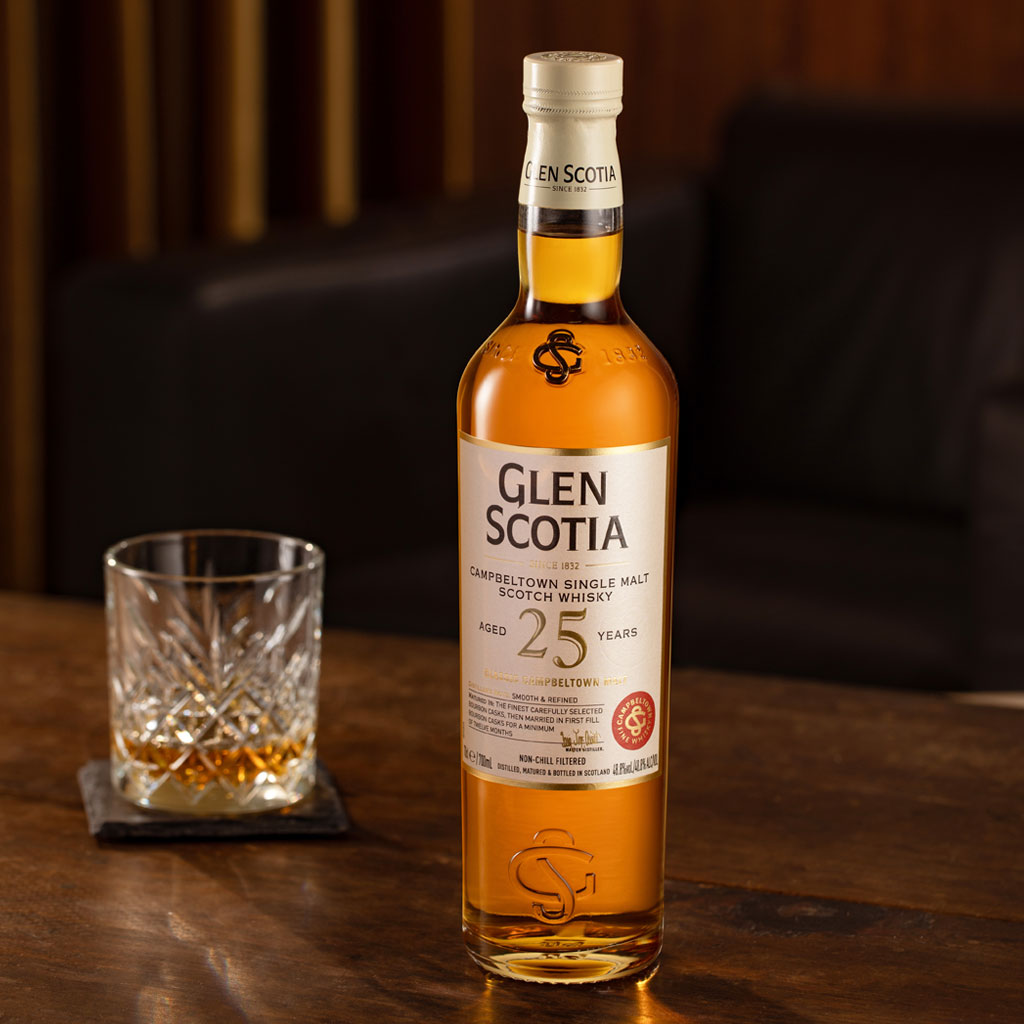 A bottle of Glen Scotia 25-year-old Campbeltown Single Malt Scotch Whisky stands on a wooden table. Beside it, a crystal glass filled with the same whisky rests on a black coaster. The background features a dark-colored couch.