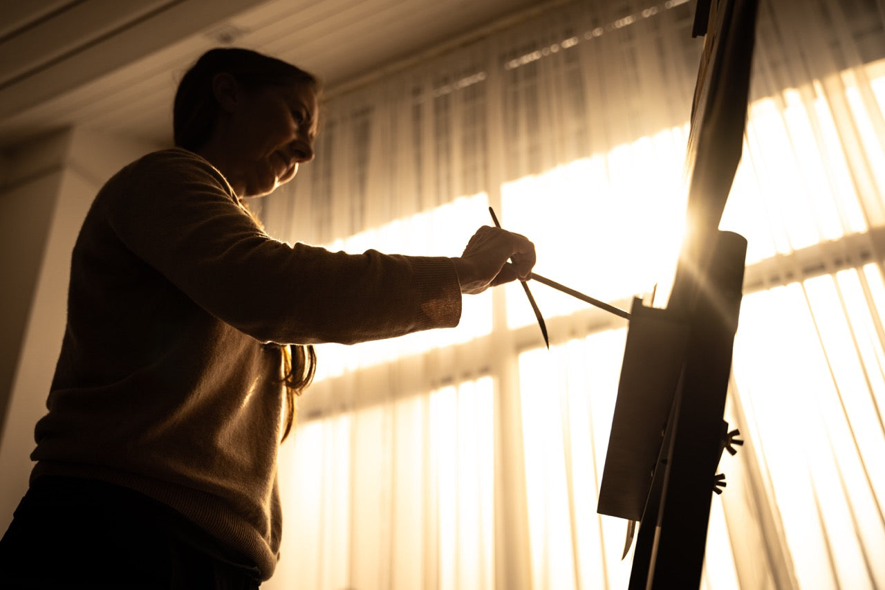 A person silhouetted against bright sunlit curtains is painting on an easel. The light creates a warm glow around them, emphasizing their focused posture and the paintbrush in hand. Art materials are visible in the foreground.