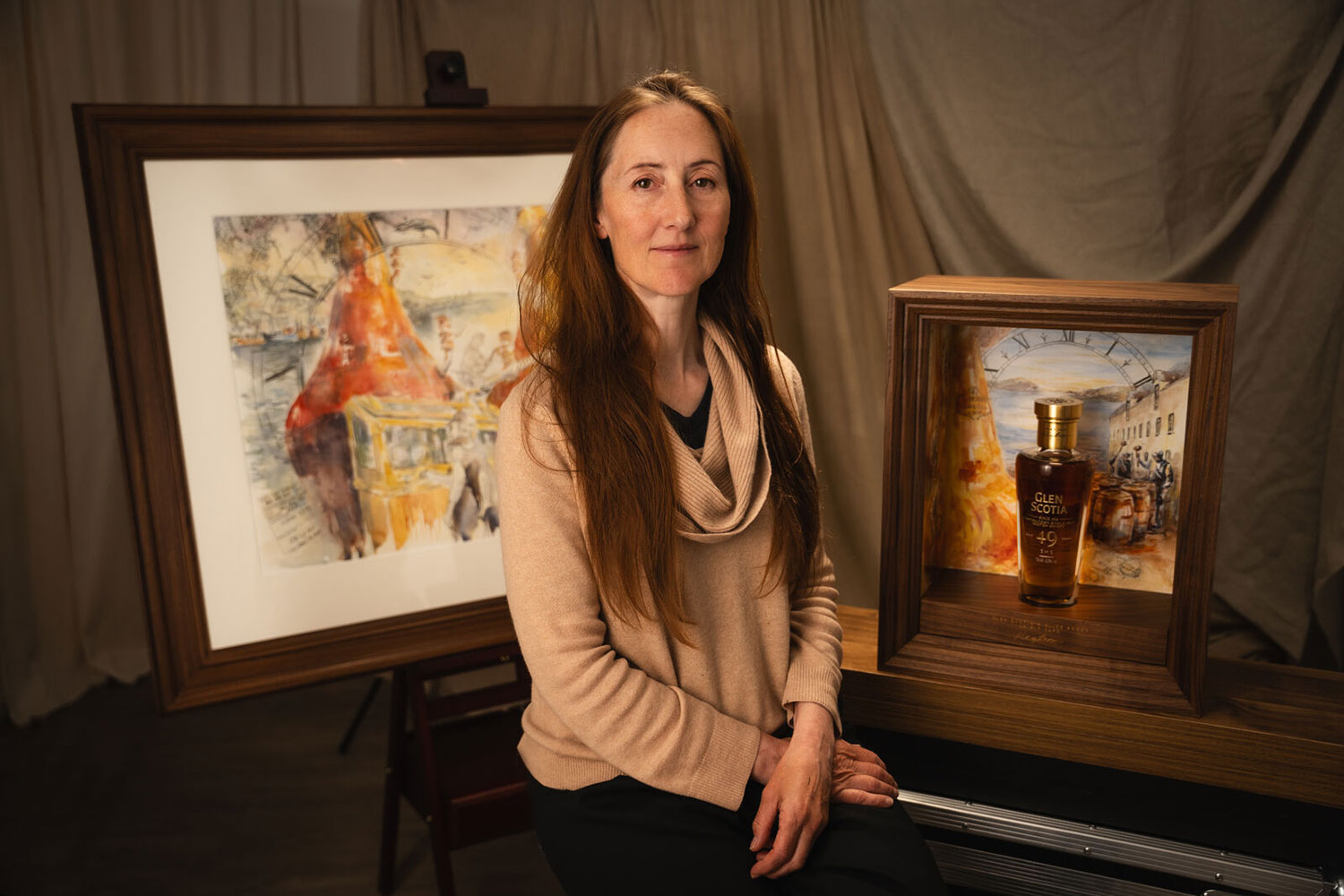 A woman with long brown hair, wearing a beige sweater, sits between two framed paintings. One painting is larger and behind her, while the other is smaller and next to her, featuring a bottle of Glen Scotia whiskey. The background includes neutral draped fabric.