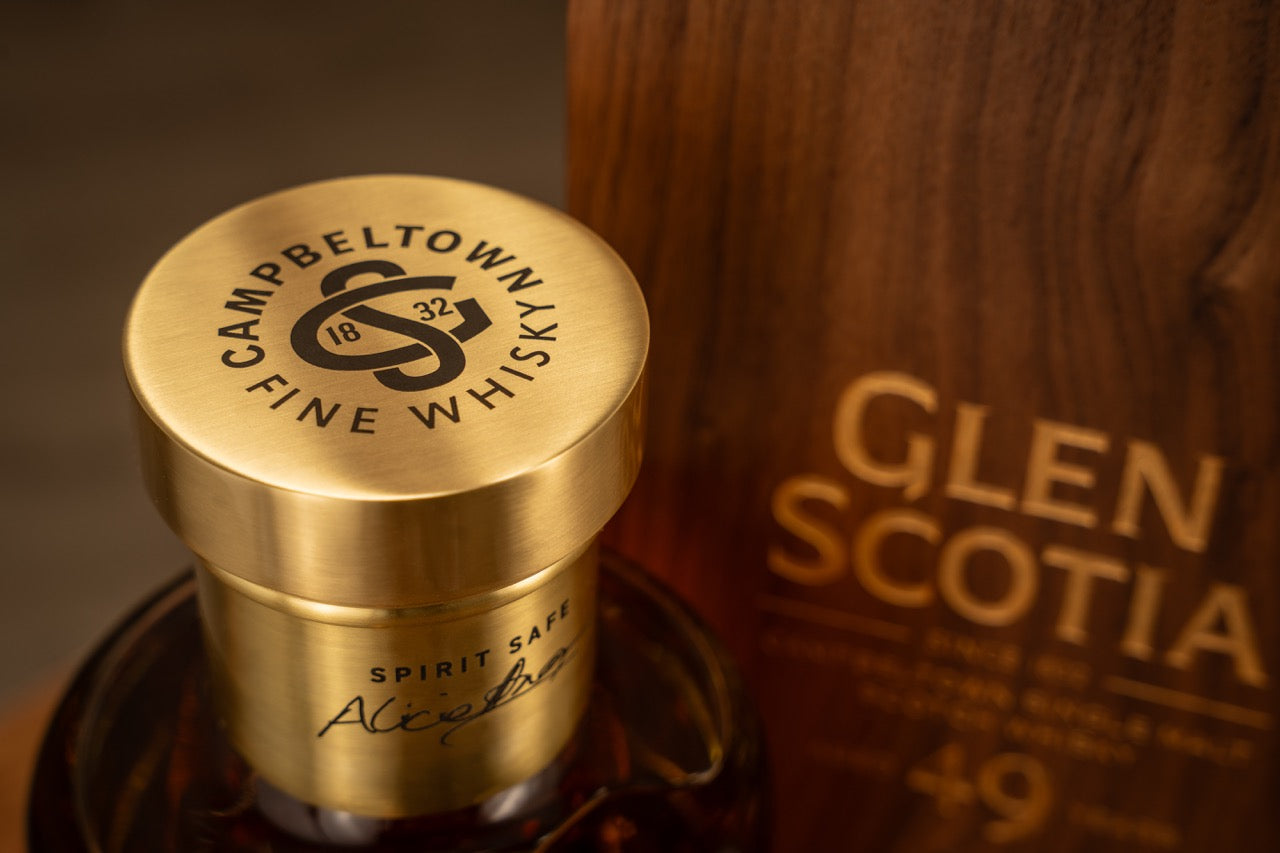 Close-up of a bottle of Campbeltown fine whisky with a gold cap bearing the brand's logo and the year 1832. To the right, a wooden box reads "Glen Scotia" with an inscription underneath. The words "SPIRIT SAFE" and a signature are visible on the bottle’s label.