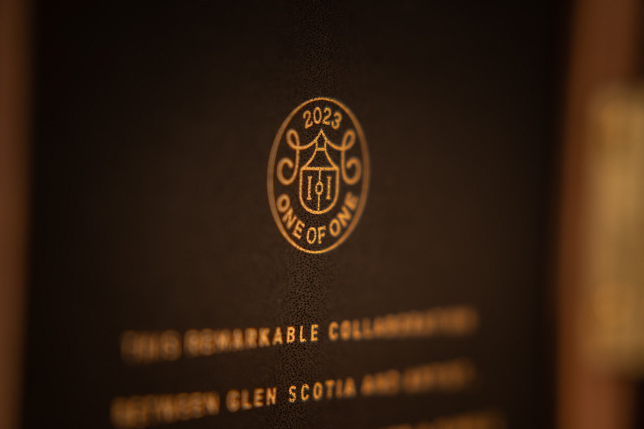 A close-up of an embossed logo on a brown surface, featuring the text "2023 ONE OF ONE" with a crest design in the center. Below the logo, partially blurred text reads, "THIS REMARKABLE COLLABORATION BETWEEN GLEN SCOTIA AND (blurred text).