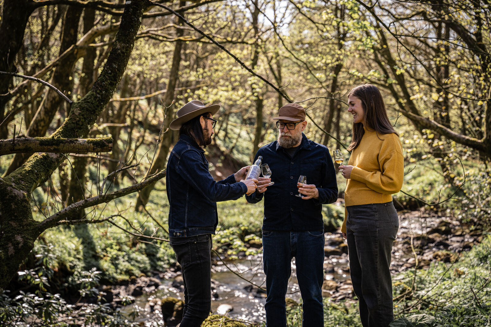 Three people stand in a forested area near a stream, sharing drinks. One person is pouring a drink into a glass held by another. The setting is lush with greenery, and they appear to be enjoying a casual outdoor gathering.