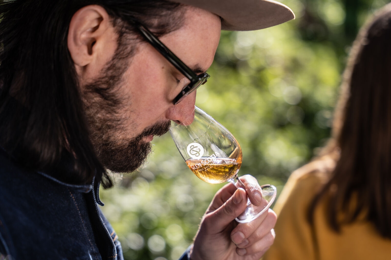 A person with long hair, wearing glasses, a hat, and a denim jacket, inhales the aroma of a drink in a wine glass marked with a logo. The background is green, suggesting an outdoor setting. Another person with long hair appears partially in the background.