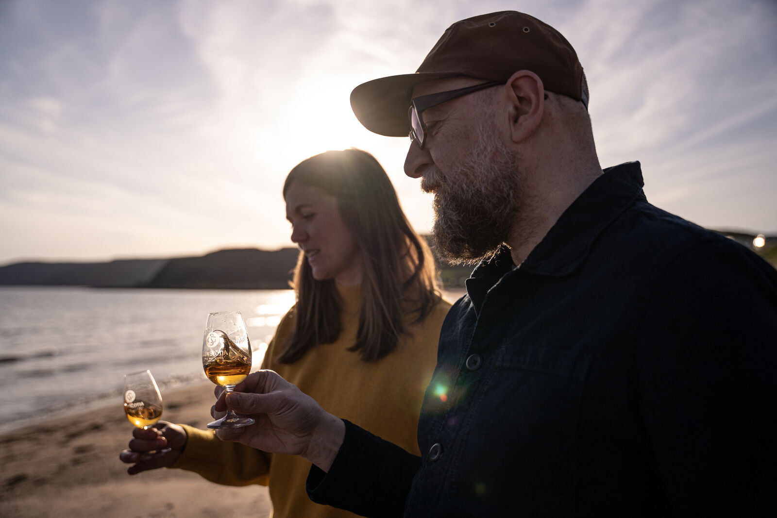 A man and woman standing on a beach, holding and examining glasses of whiskey against the setting sun. The man wears glasses and a cap, while the woman wears a yellow sweater. The calm sea and hills are visible in the background.