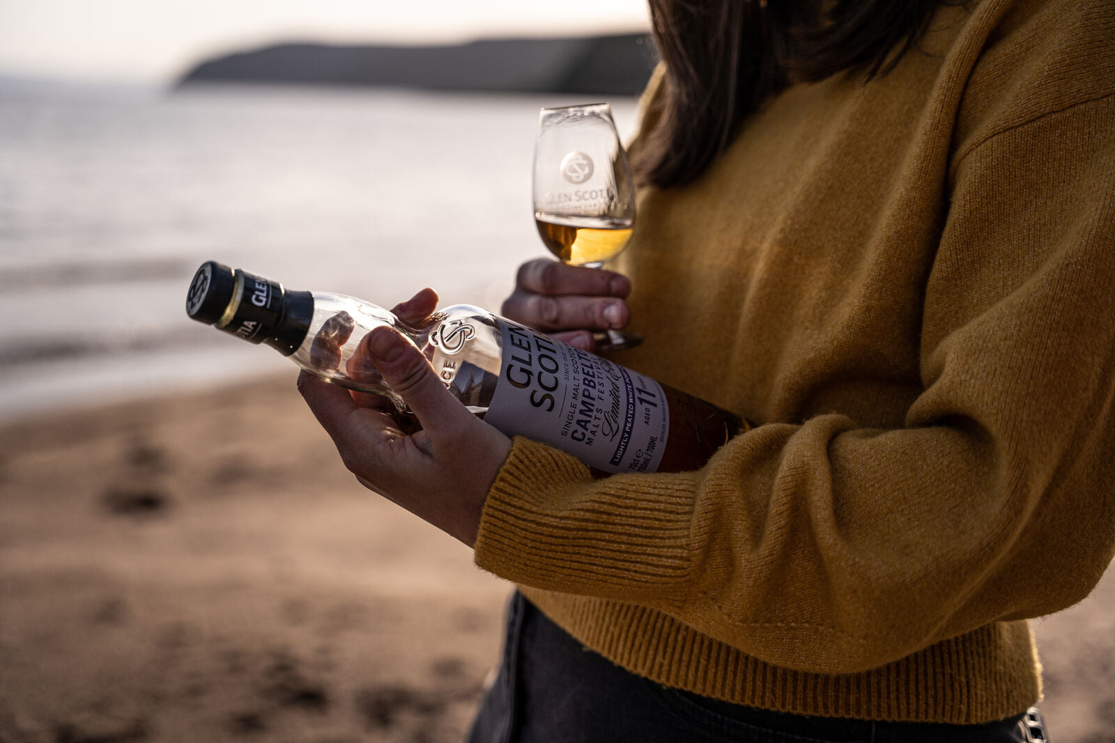 A person in a yellow sweater holds a bottle of Glen Scotia Scotch whisky in one hand and a glass of whisky in the other while standing on a beach with a blurred shoreline and hills in the background. The scene suggests a relaxing, scenic whisky-tasting moment.