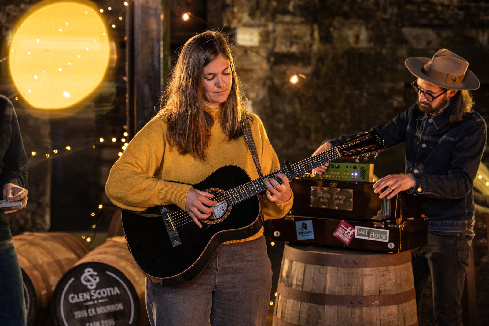 A woman in a yellow sweater plays an acoustic guitar in a dimly lit setting with string lights and a large barrel marked "Glen Scotia" nearby. A man beside her, wearing a hat and glasses, adjusts equipment on another barrel. The atmosphere is cozy and intimate.