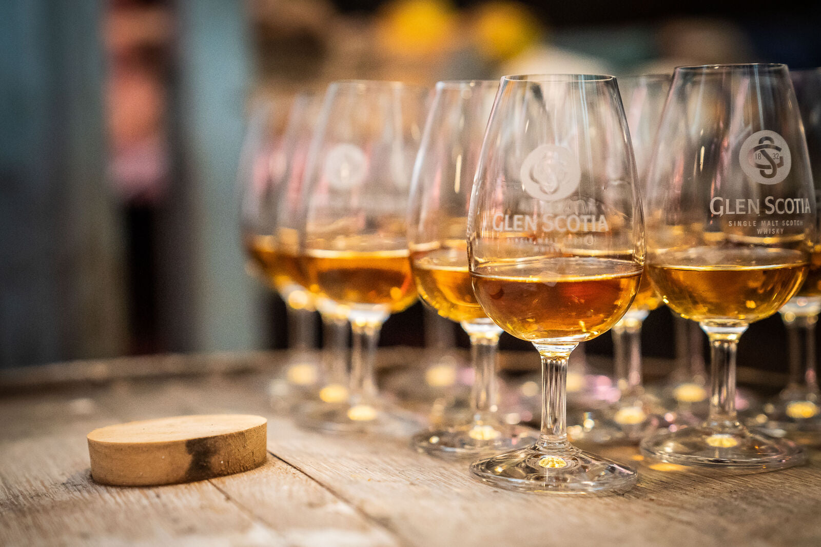 Close-up of nine glasses of Glen Scotia single malt scotch whisky on a wooden surface, arranged in a row with an extra wooden coaster on the left. The liquid in the glasses is a rich amber color, and the background is softly blurred, creating a warm ambiance.
