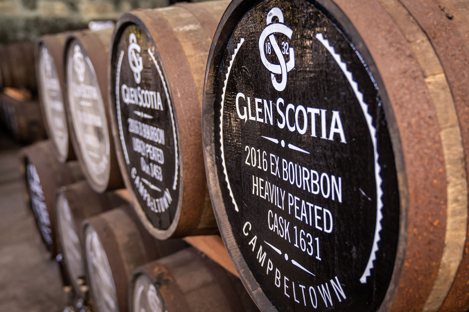 Close-up of several stacked wooden barrels. The front barrel is labeled "Glen Scotia 2016 Ex Bourbon Heavily Peated Cask 1631 Campbeltown." The barrels are stored in a dimly lit area, possibly a distillery or a cellar.
