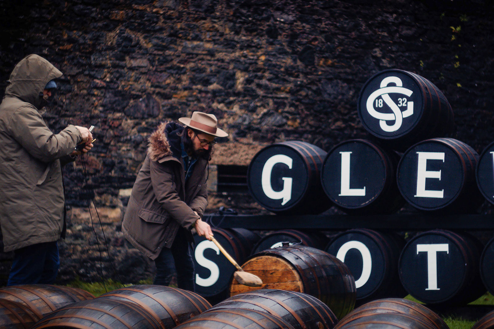 Two individuals are outdoors, with one wearing a hat and using a tool to work on a barrel, while the other observes or records. They are surrounded by stacked wooden barrels labeled with white letters forming "GLEN SCOTIA," against a stone wall. The scene suggests a distillery.