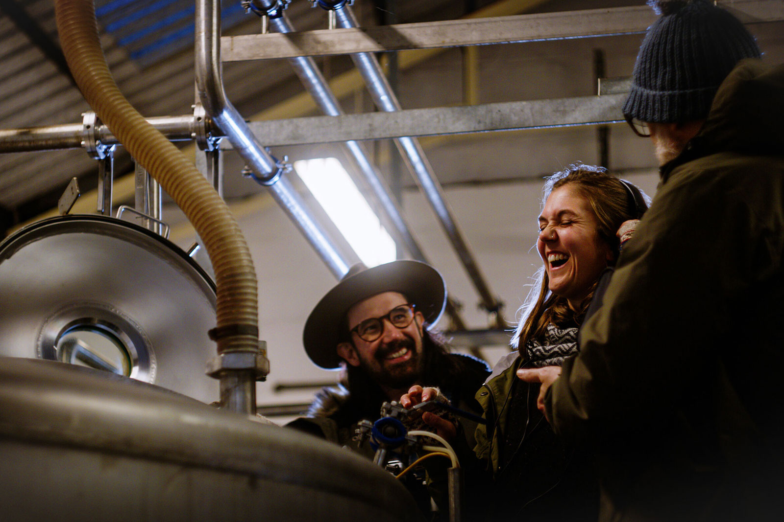 Three people in a brewery laugh together near large metal brewing equipment. The person in the foreground wears a hat and glasses, the one in the center with long hair laughs heartily, and the third person wears a beanie, and has a white beard.