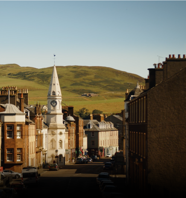 A picturesque town with a prominent white clock tower stands in the center, flanked by buildings on both sides. Green rolling hills rise in the background under a clear blue sky. Vehicles and a few people are visible on the street.