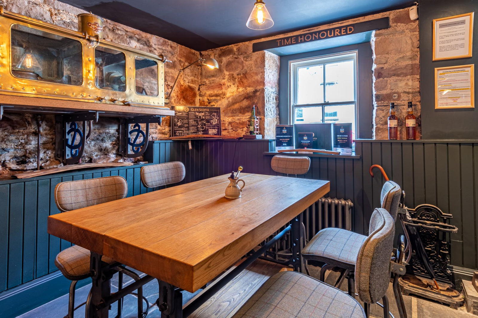 A cozy dining area with a wooden table surrounded by upholstered chairs. The walls are a mix of exposed brick and dark green wainscoting. A shelf with vintage items is illuminated above one wall. A window lets in natural light, and framed certificates hang nearby.
