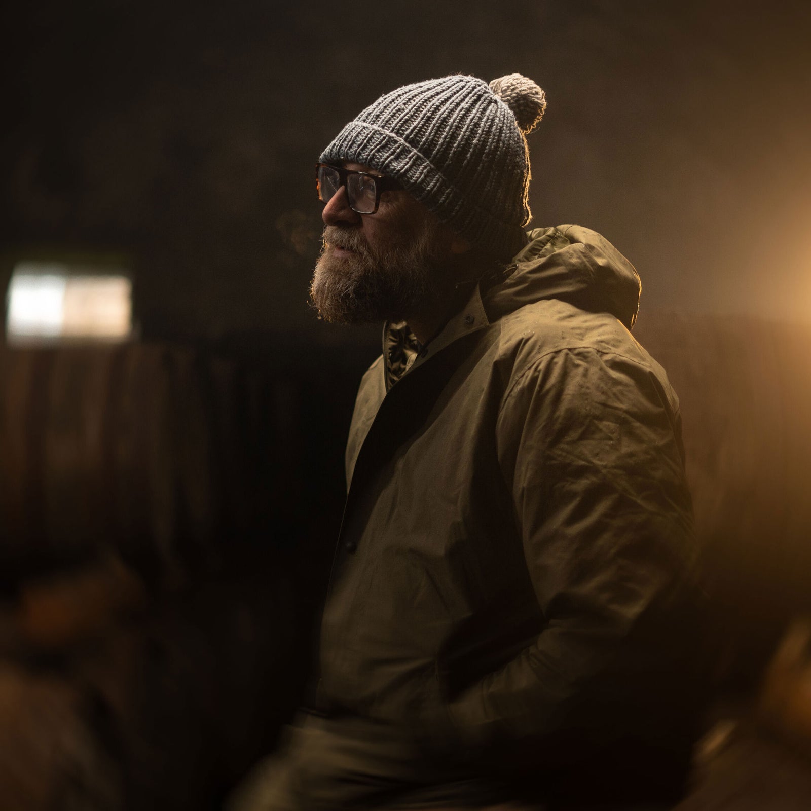A person with a beard and glasses wears a knitted beanie and a rain jacket. They are looking to the side in a dimly lit environment, with soft, warm light illuminating their profile from the right. The background is dark and out of focus.