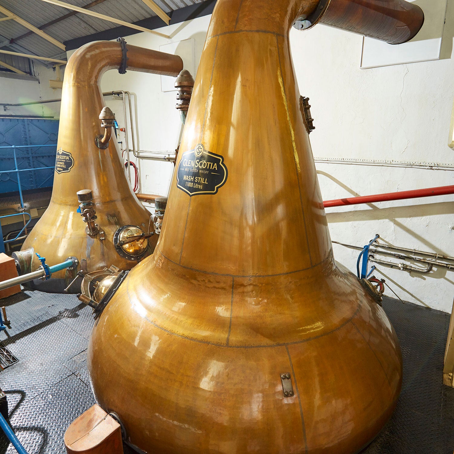 A photo of copper pot stills in a distillery. The stills are labeled "Glen Scotia Wash Still." The room has industrial lighting, piping, and metallic flooring. The copper stills have a polished and worn finish, typical of whisky distillation equipment.