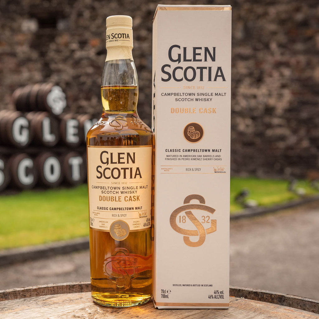 A bottle of Glen Scotia Campbeltown Single Malt Scotch Whisky Double Cask stands next to its matching cream-colored box. The box and bottle display the whisky's details, including its age and origin. A blurred outdoor stone wall forms the background.