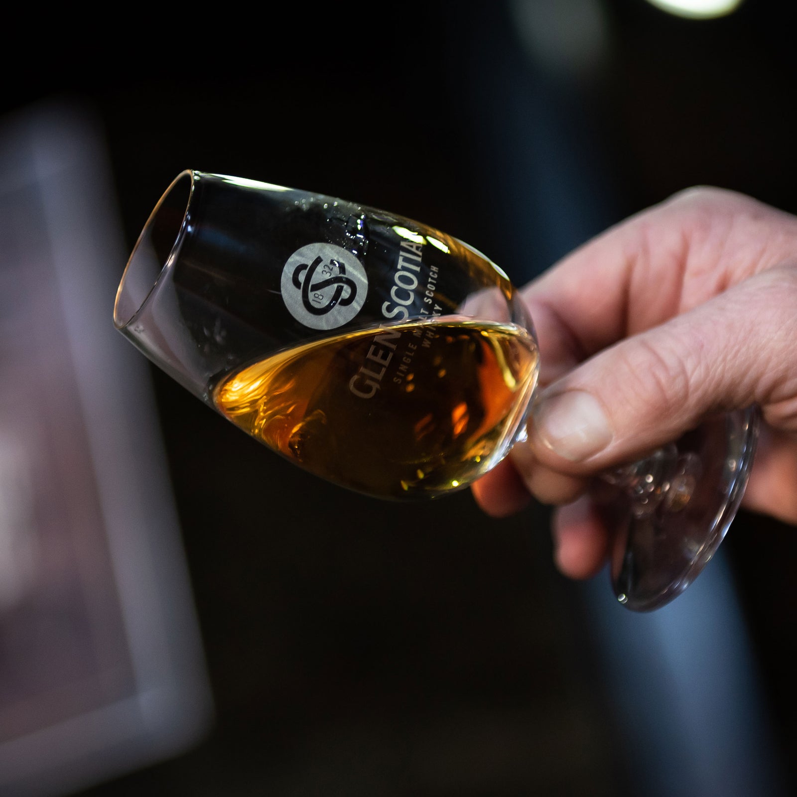 A hand holding a tulip-shaped glass with "Glen Scotia" logo on it, filled with a rich amber-colored whisky. The background is blurred, focusing attention on the glass and the whisky's color.