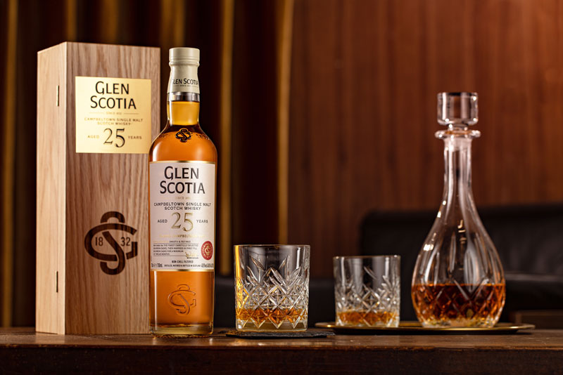 A bottle of Glen Scotia 25-year-old single malt whiskey stands next to its wooden box. Two crystal glasses filled with whiskey sit in front of the bottle, and a crystal decanter partially filled with whiskey stands beside the glasses on a wooden surface.