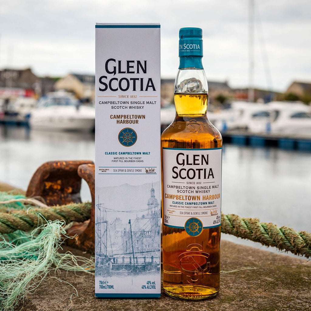 A bottle of Glen Scotia Campbeltown Harbour single malt Scotch whisky stands next to its box on a stone surface with fishing nets in the foreground. The background shows a marina with boats, adding a nautical theme reflective of the whisky's coastal origin.