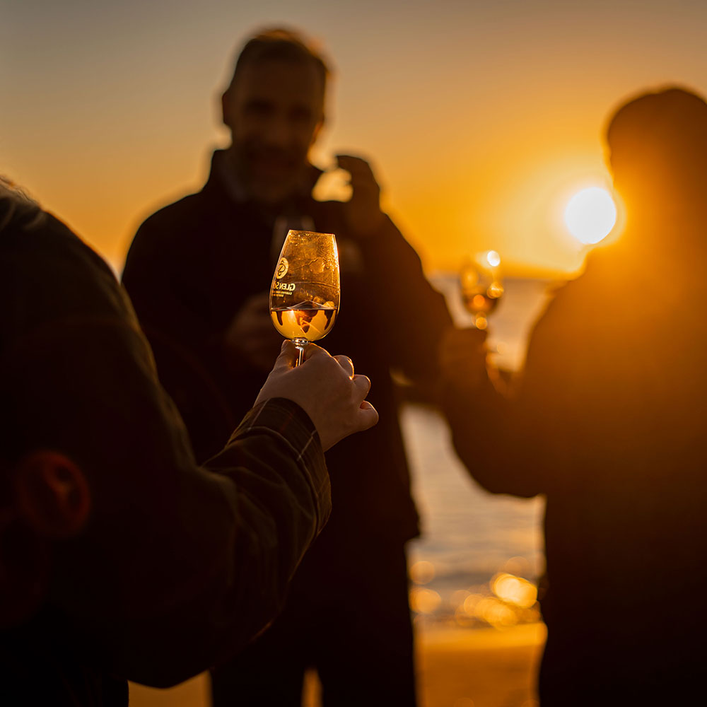 People silhouetted against a sunset, holding glasses of amber liquid, possibly whiskey or wine. The setting sun casts a warm, golden glow over the scene, with the ocean visible in the background. The focus is on the raised glass in the foreground.