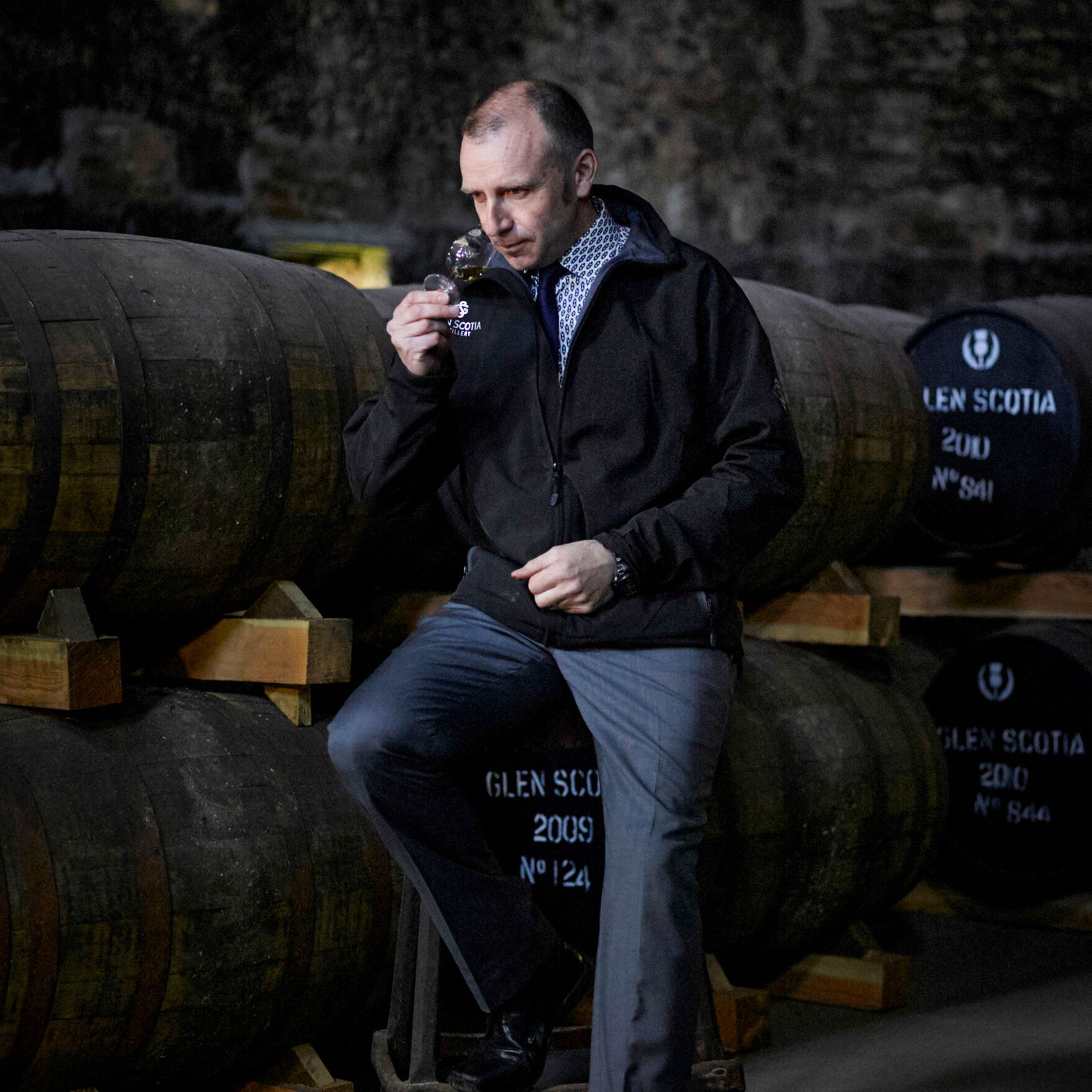 A man in a black jacket and gray pants is sitting on stacked wooden barrels in a dimly lit cellar. He is holding a glass and smelling its contents, with more large barrels marked "Glen Scotia" visible in the background.
