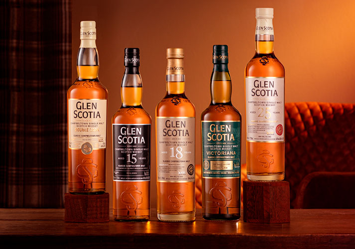 Five bottles of Glen Scotia Scotch whisky are lined up on a wooden surface. The labels display various age statements and expressions: Double Cask, 15 years, 18 years, Victoriana, and 25 years. The background features dark wood and a plaid pattern.
