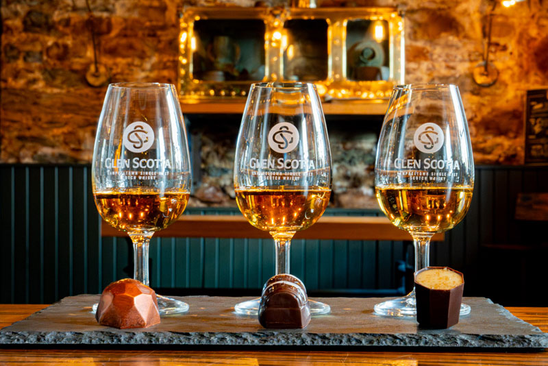 Three Glen Scotia branded glasses filled with whisky are arranged in a row on a slate tray. Each glass is paired with a distinct chocolate piece in front of it. The background features a rustic wood and stone interior of a presumably whisky tasting room.