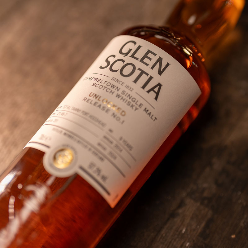 A bottle of Glen Scotia's Unlocked #1, a 9 Year Old Single Malt Scotch whisky, rests on a wooden surface, proudly displaying its Exclusive Cask edition. The label highlights "Campbeltown Single Malt" and "Unpeated Release." Inside, the warm amber liquid reveals an enticing touch of Tawny Port casks' influence.
