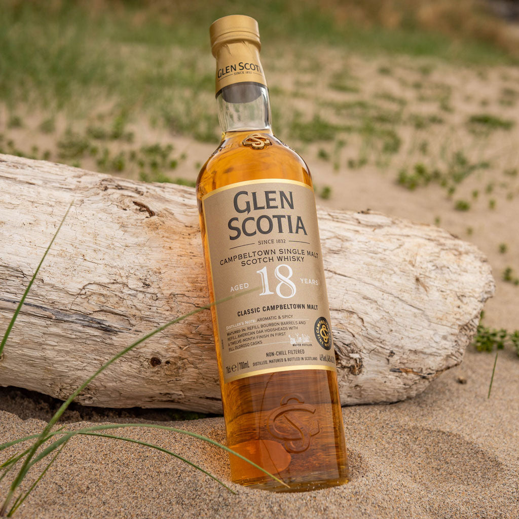 A bottle of Glen Scotia 18-year-old Campbeltown single malt Scotch whisky lies on sandy ground, leaning against a weathered piece of driftwood. The whisky bottle features a gold cap and a beige label with black and gold text.