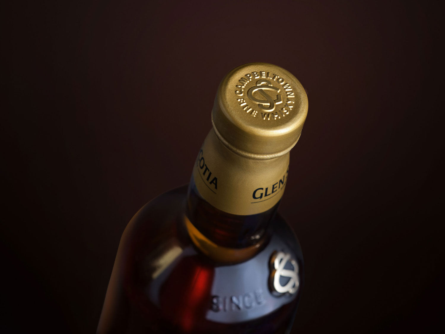 Close-up of the neck and gold-colored cap of a bottle of Glen Scotia whisky. The cap is embossed with the text "GLEN CAMPBELTOWN WHISKY". The bottle is dark brown, with some of the liquid visible. The background is dark and out of focus.