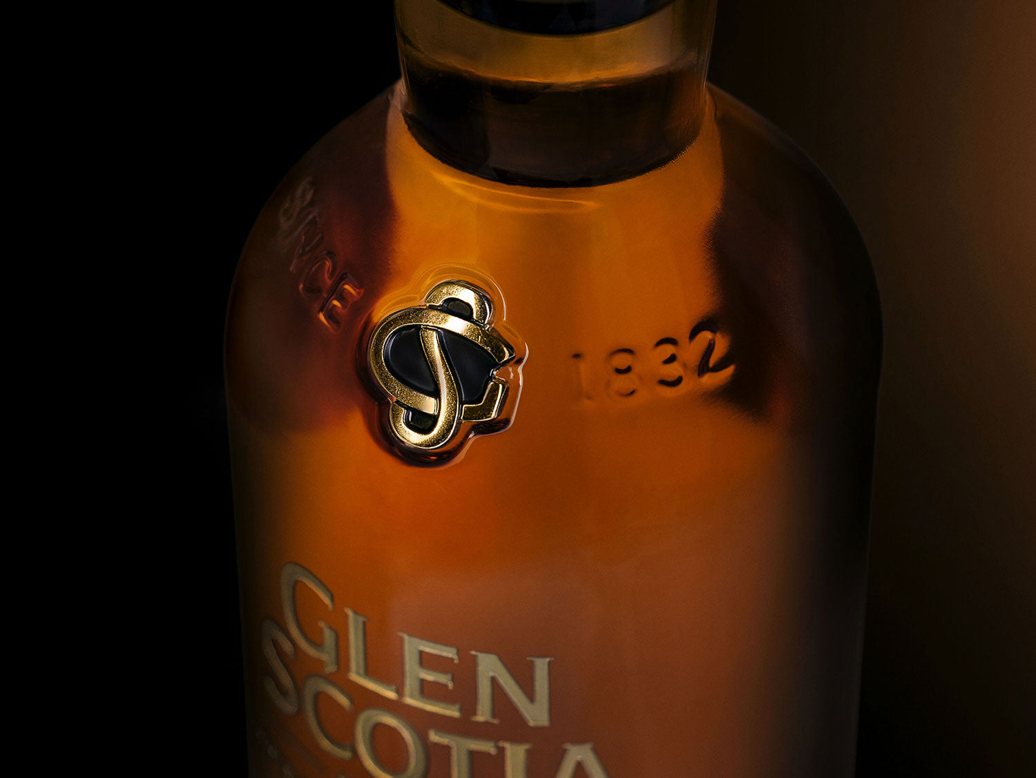 Close-up of the neck and upper label of a Glen Scotia whiskey bottle. The bottle's amber liquid is visible through the glass, showcasing an embossed date "1832" and a golden "S" emblem. The brand name "Glen Scotia" is partially visible on the bottle.