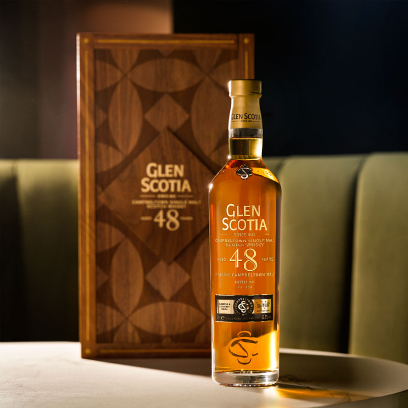 A bottle of Glen Scotia 48-year-old single malt Scotch whisky is displayed on a table in front of a wooden box with the same branding. The bottle features a gold cap and label. The background is dimly lit, focusing attention on the bottle and box.