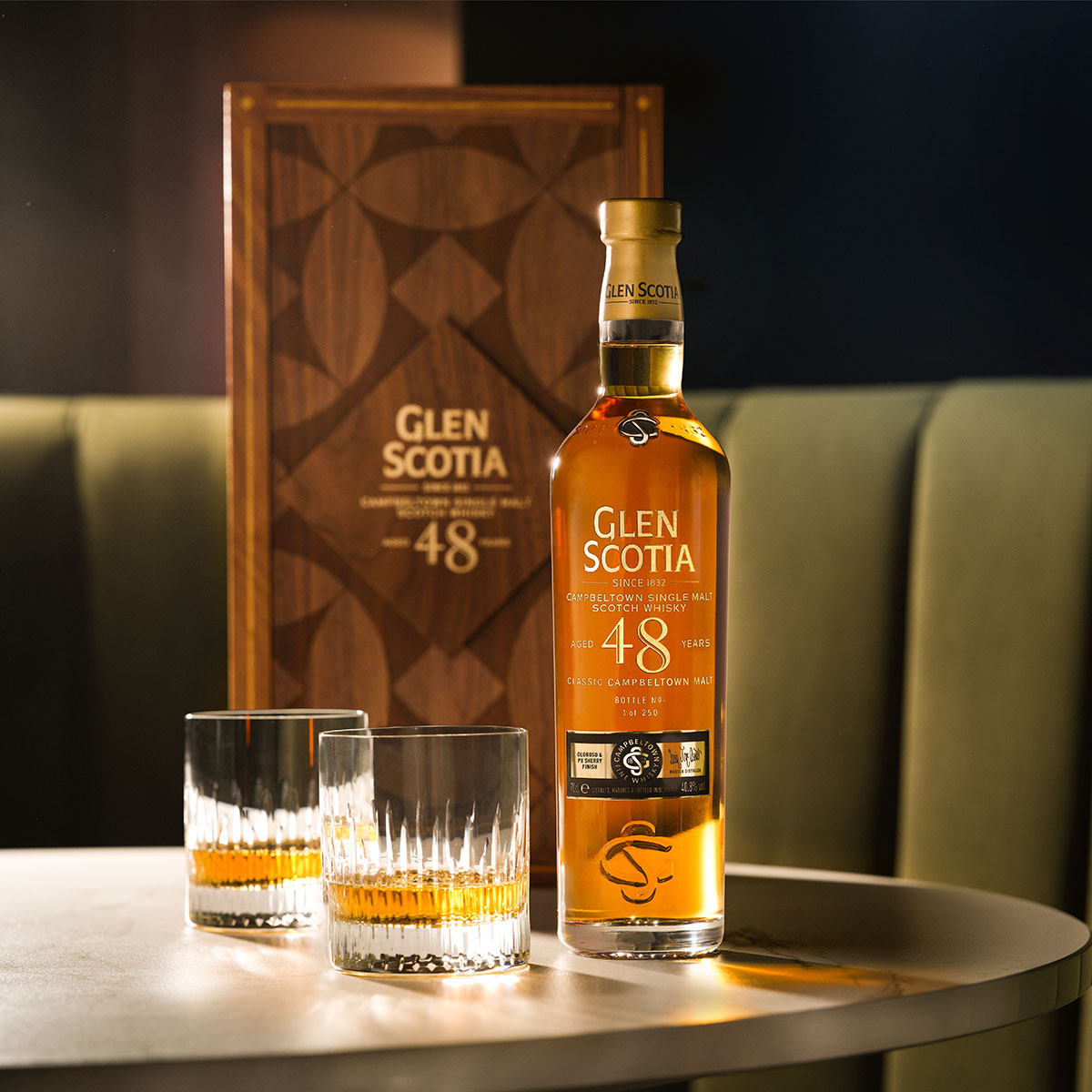 A bottle of Glen Scotia 48-year-old single malt scotch whisky is placed on a table with two filled whisky glasses. The bottle is positioned in front of its decorative case, both sitting on a round, light-colored table against a green cushioned background.
