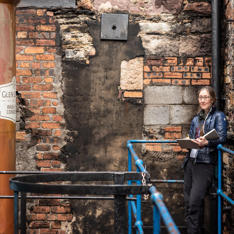 A person with glasses stands on a blue metal staircase beside an aged brick wall, holding a notebook and pen. A large metallic cylinder with "GLEN" partially visible is to the left. The scene looks like an industrial or rugged historical site.
