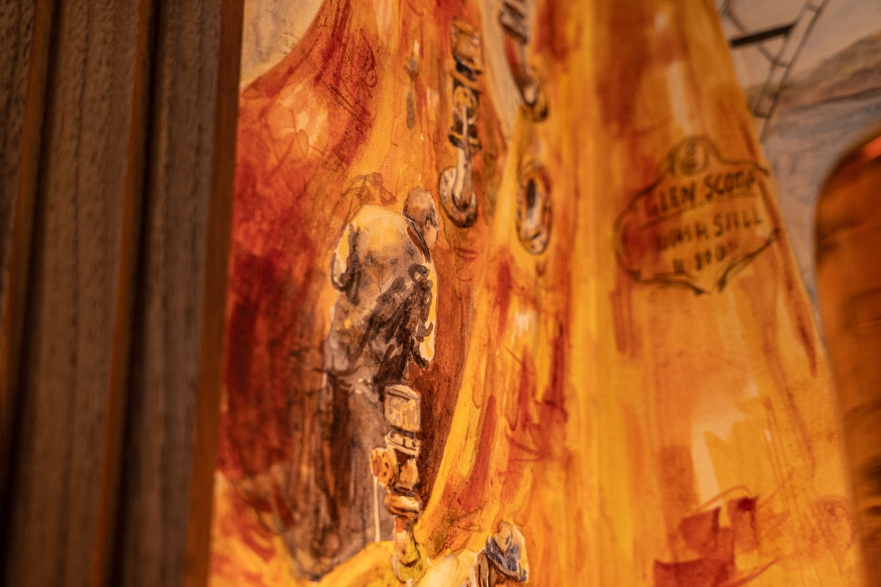 Close-up of a painting showing an industrial scene with workers wearing helmets and gear amidst machinery. The predominant colors are warm tones of orange and yellow, creating a vibrant atmosphere. A small sign with partially readable text is visible on the right side.