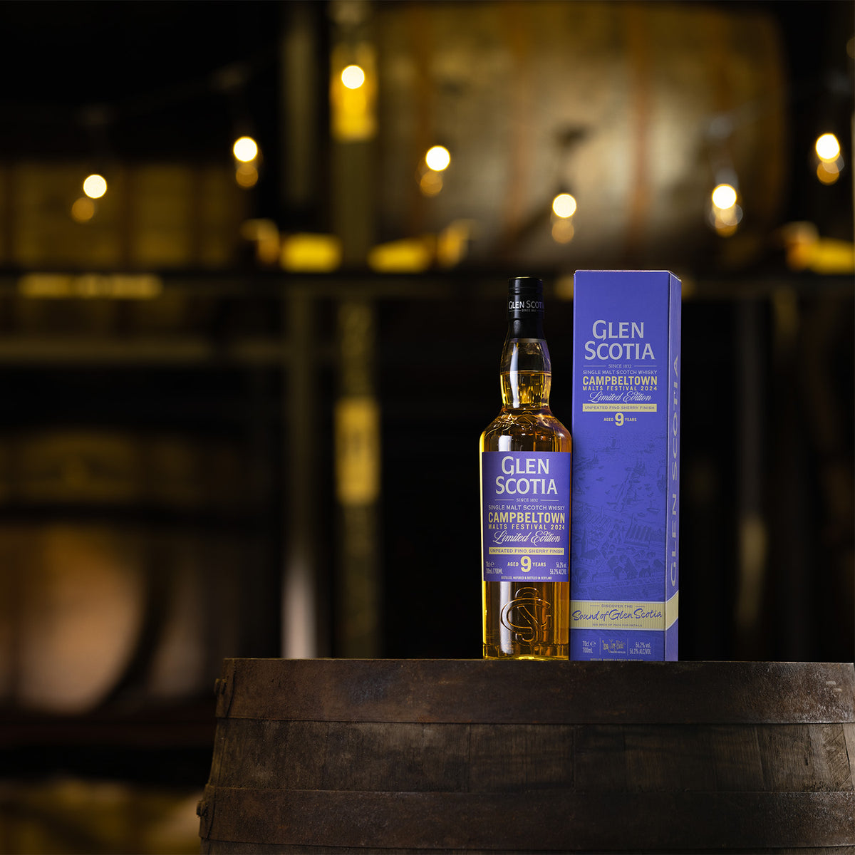 A bottle of Glen Scotia Campbeltown Malts Festival Edition 2024 stands next to its blue and yellow packaging on a wooden barrel. The background features dimly lit wooden barrels and hanging lights, creating a cozy and rustic atmosphere, perfect for those seeking unique whisky gifts.