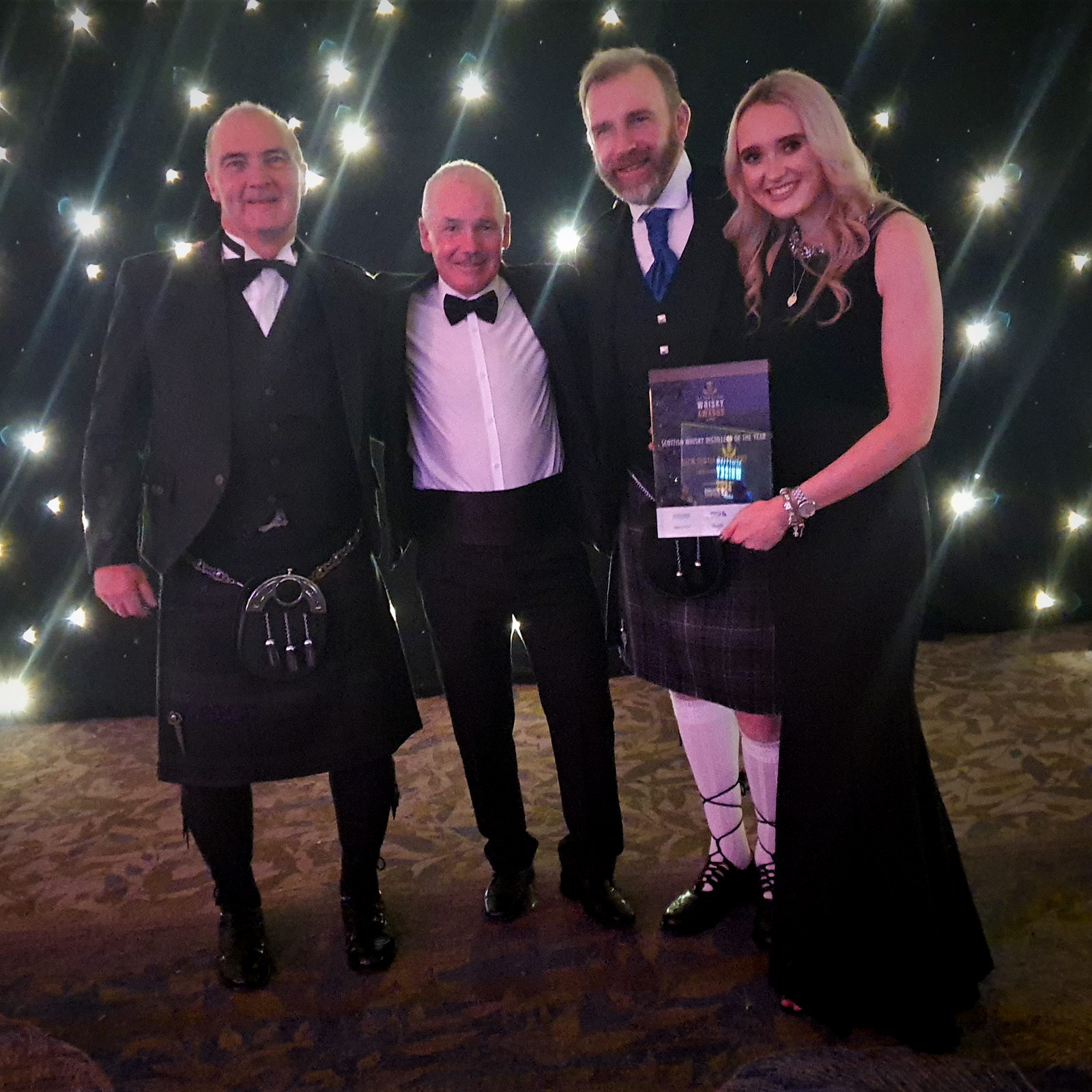 Four people pose together for a photo at an event. Three men, two in kilts and one in a tuxedo, stand next to a woman wearing a black dress. They are in front of a backdrop with twinkling lights. One man is holding a plaque or award.