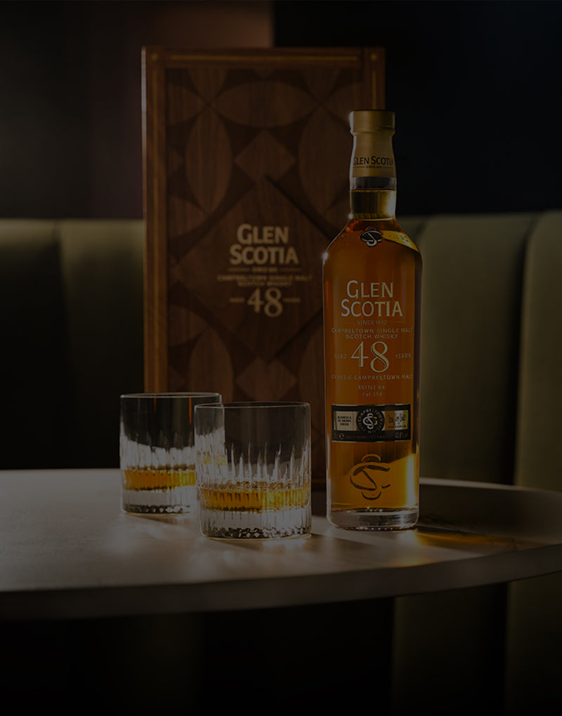A bottle of Glen Scotia 48-year-old single malt Scotch whisky stands on a table beside two crystal glasses filled with whisky. Behind the table is the wooden whisky case, displaying its name and age. The background is dark and slightly out of focus.