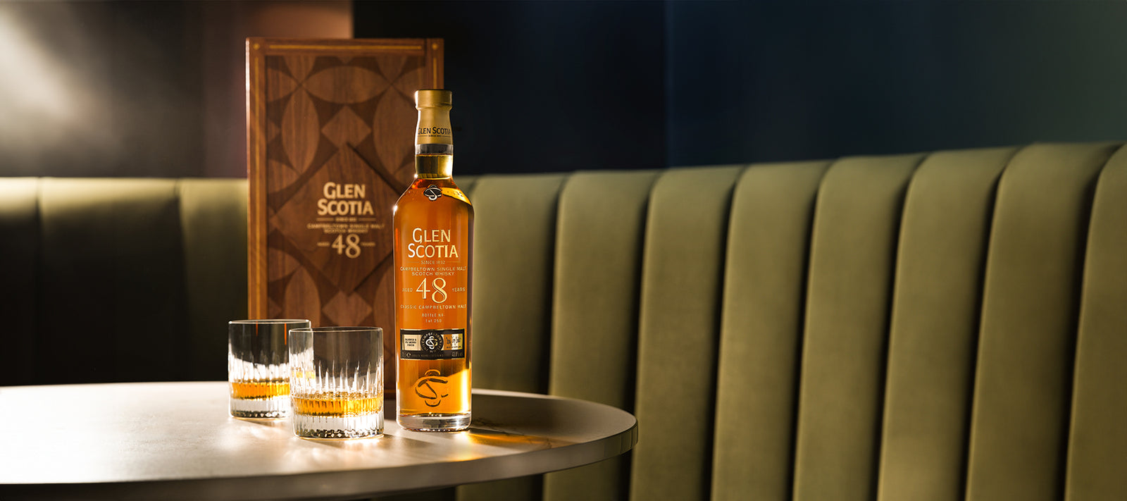 A bottle of Glen Scotia 48-year-old whiskey sits on a table beside two filled whiskey glasses. The table is positioned against a green upholstered booth. The whiskey box is also on the table behind the bottle. The setting has a dark, elegant ambiance.