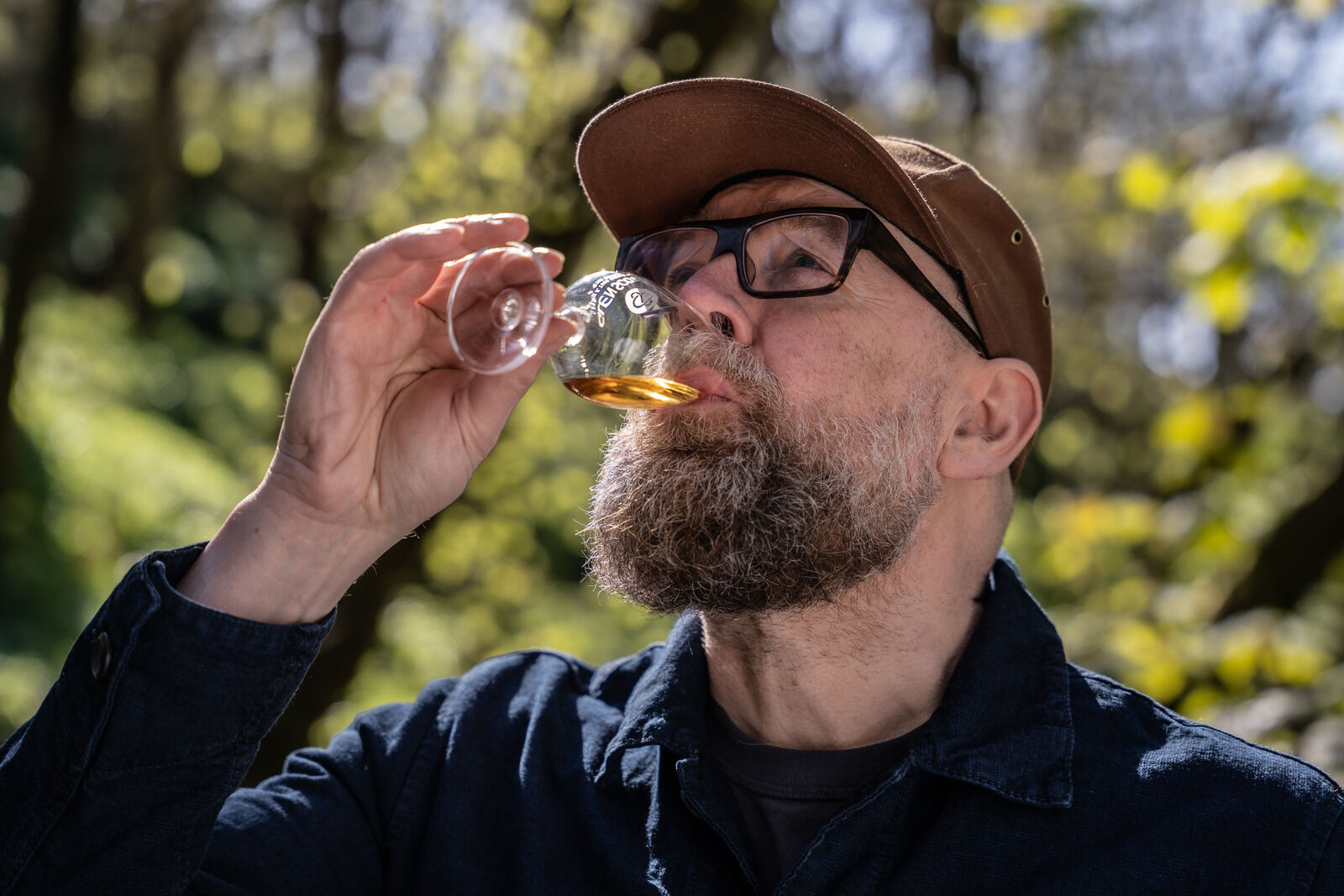 A man with a beard, wearing a brown cap and black glasses, is outdoors, taking a sip from a small glass of amber-colored liquid. He is dressed in a dark blue jacket. Trees with green leaves are visible in the blurred background.