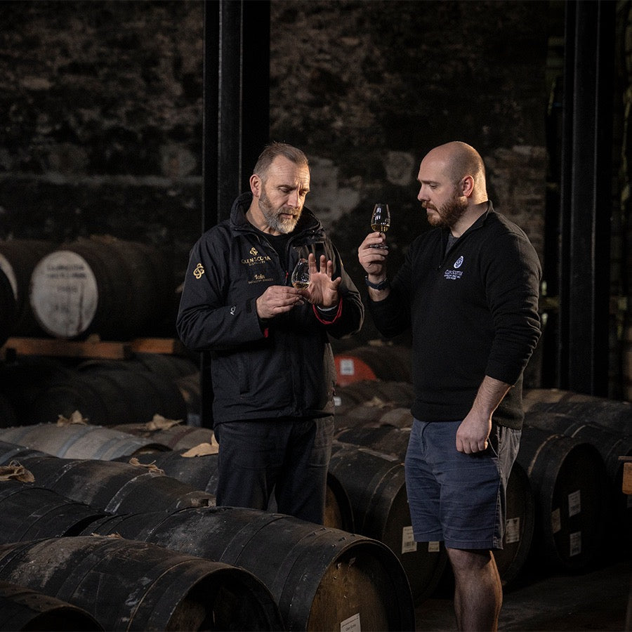 Two men are standing in a dimly lit cellar, surrounded by wooden barrels, each holding a glass of dark liquid. The man on the left, gesturing with his hand, is in mid-conversation while the man on the right listens attentively.