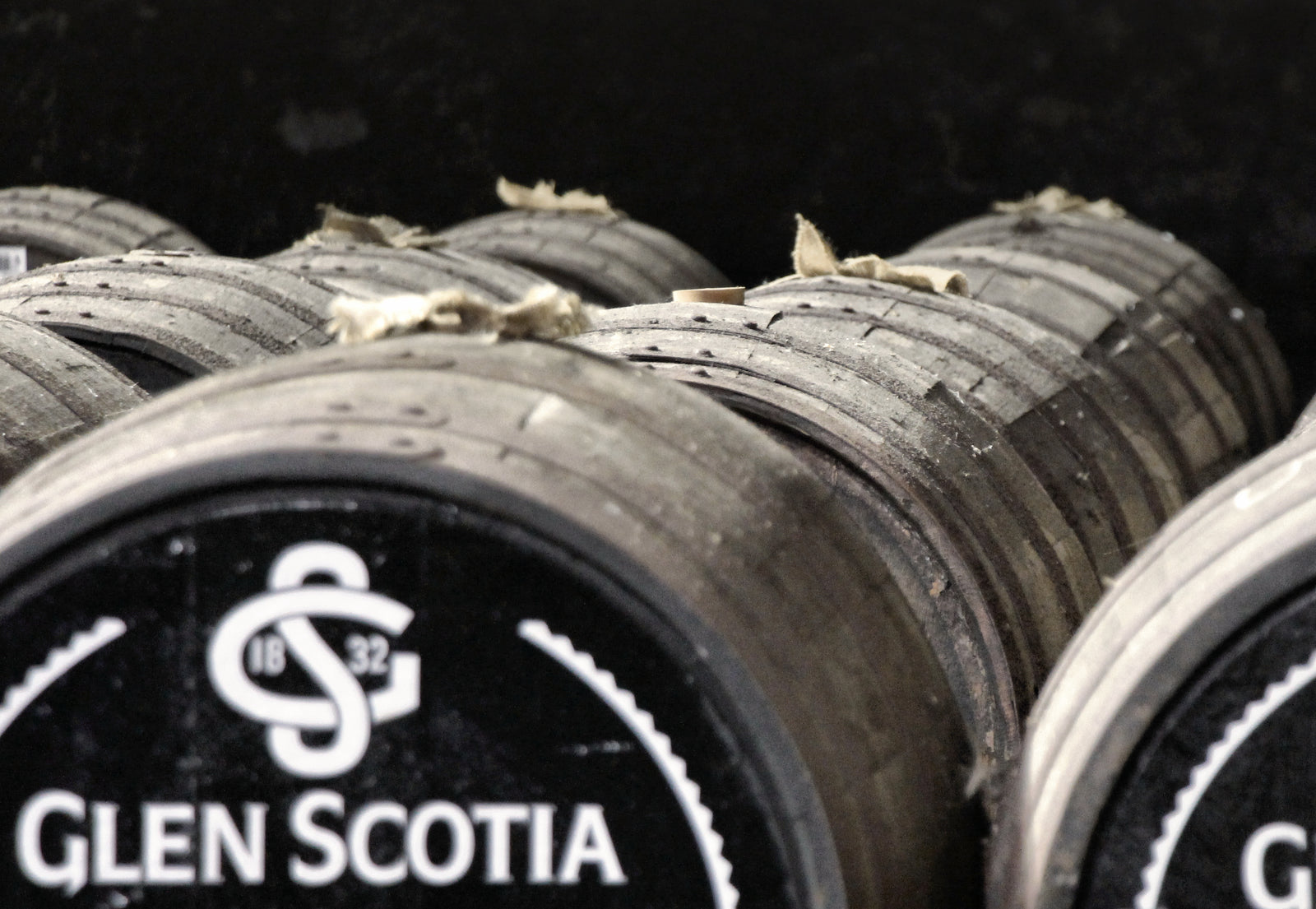 A stack of wooden barrels is shown in a dimly lit storage area. The barrels have the words "Glen Scotia" and the number "1832" printed on the ends, which indicates they are used for aging spirits. The tops of the barrels appear dusty and old.