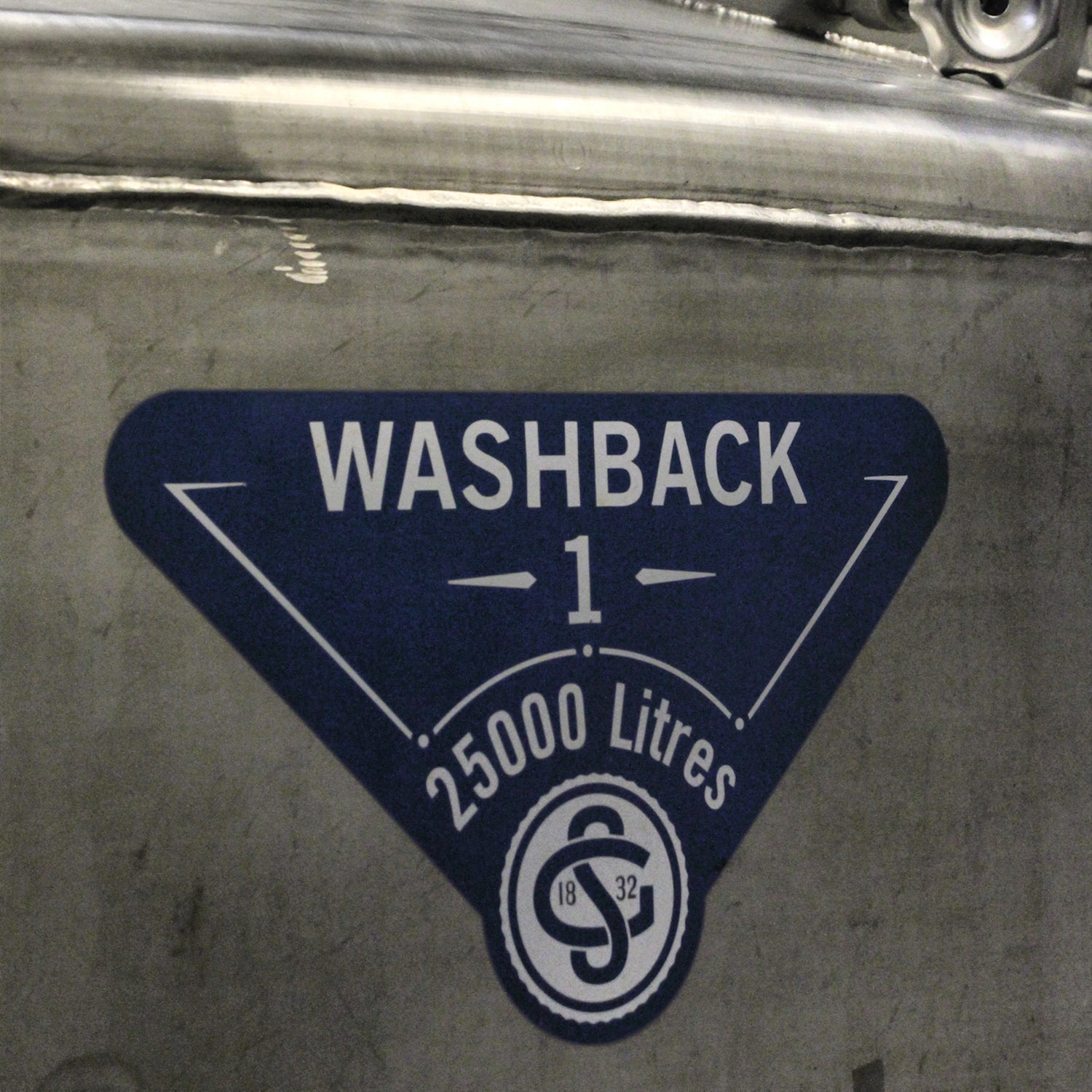 A metallic surface with a triangular blue label featuring the text "WASHBACK 1" and "25000 Litres." Below the text, there is a logo with the numbers "18" and "32.