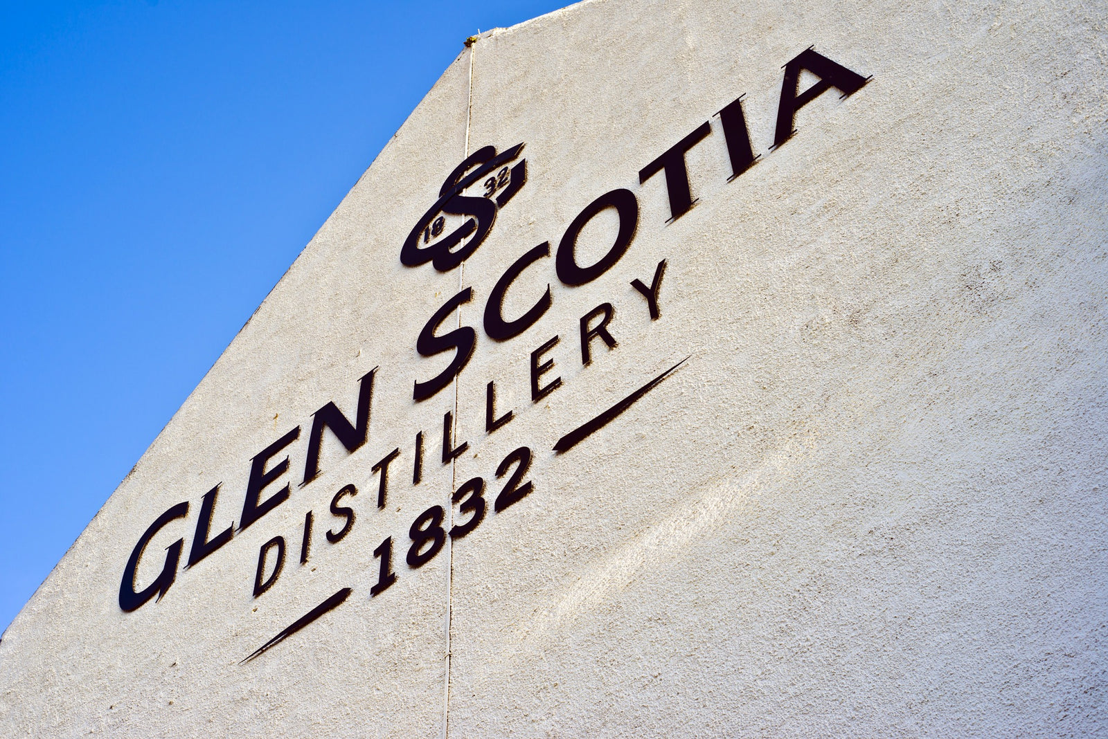 A close-up of the exterior wall of Glen Scotia Distillery. The wall features the distillery's name, logo, and founding year "1832" prominently displayed in large, bold lettering against a clear blue sky. The surface of the wall is textured and cream-colored.