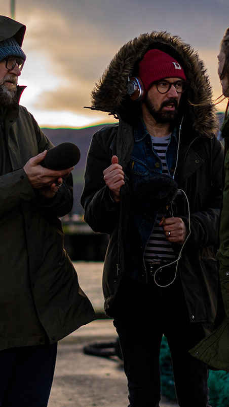Three people are outdoors in a cold, overcast setting. The person in the center, wearing a red beanie and headphones, holds a microphone covered with a windscreen. The individuals appear to be in conversation, dressed in warm clothing with fur-hooded jackets.