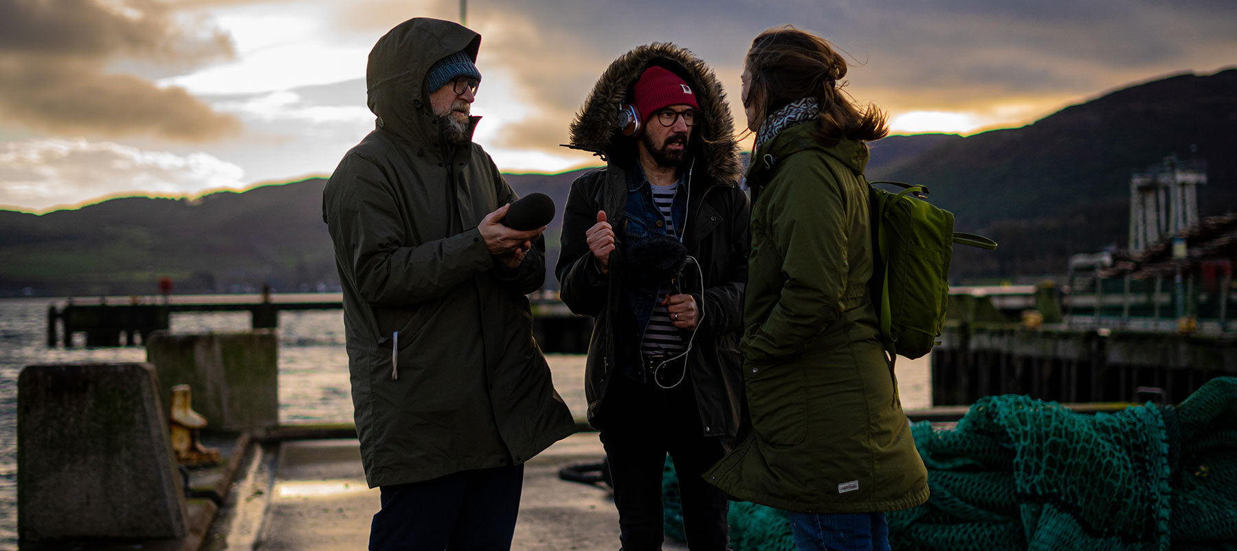 Three people in warm clothing stand on a pier in conversation during sunset. The person in the center wears a red beanie, hooded parka, and glasses, holding a microphone. The person on the left holds a black object, and the person on the right carries a green backpack.