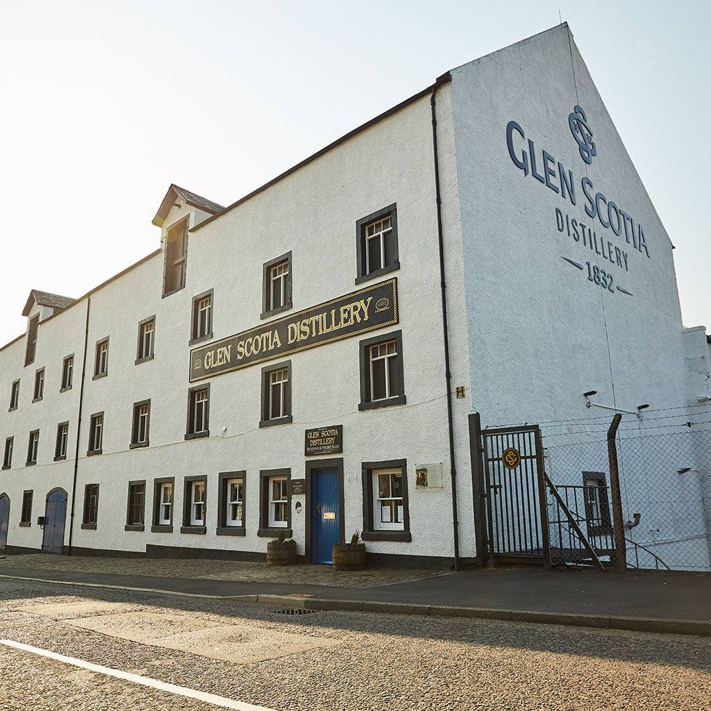 A large, white, multi-story building with "Glen Scotia Distillery" painted on it. The facade features several windows and a sign above the main entrance door, also saying "Glen Scotia Distillery". A fenced gate is visible on the right side of the building.