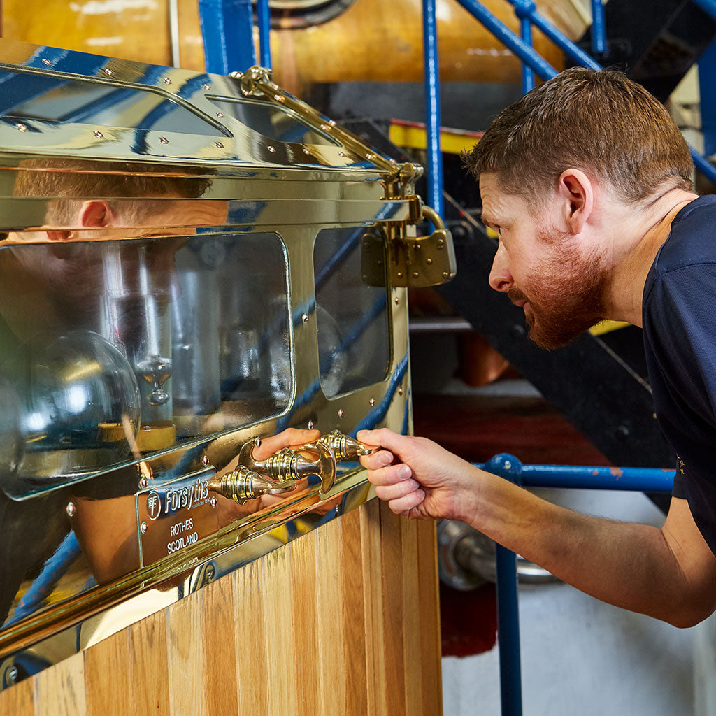 A person with short hair and a beard closely inspects a polished brass and wood distillation apparatus labeled “Forbes, Rothes, Scotland.” The setting appears industrial, with blue metal railings and machinery in the background.