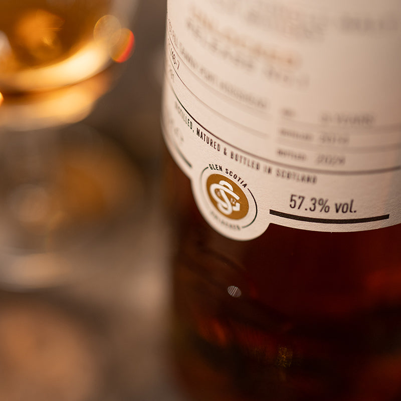 Close-up of a whiskey bottle showing an alcohol content of 57.3%, with a glass of Unlocked #1, a 9-Year-Old Glen Scotia Single Malt, in the blurred background. The label reveals it was distilled and bottled in Scotland, aged in exclusive casks for enhanced richness.